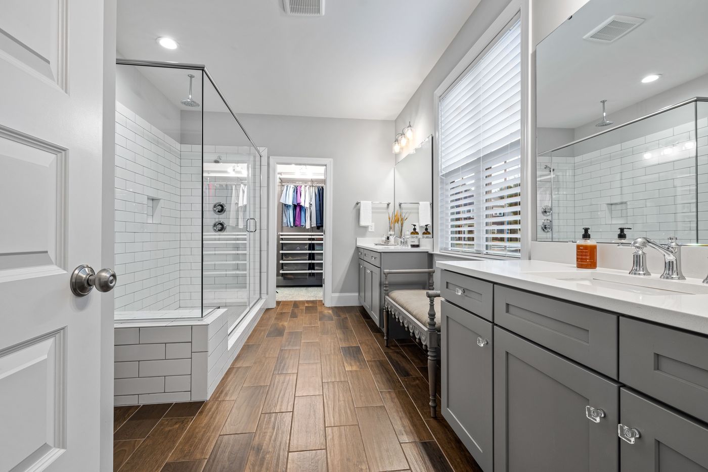 Primary bath with gray vanities, glass shower, and a walk-in closet beyond